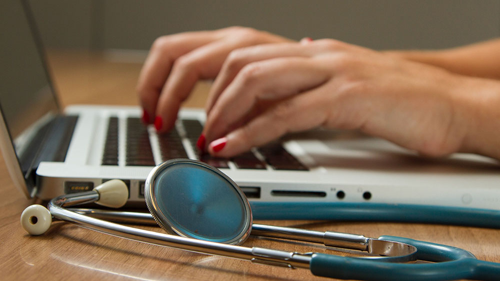 A lady's hands typing on a laptop with a stethoscope next to the computer on the desk